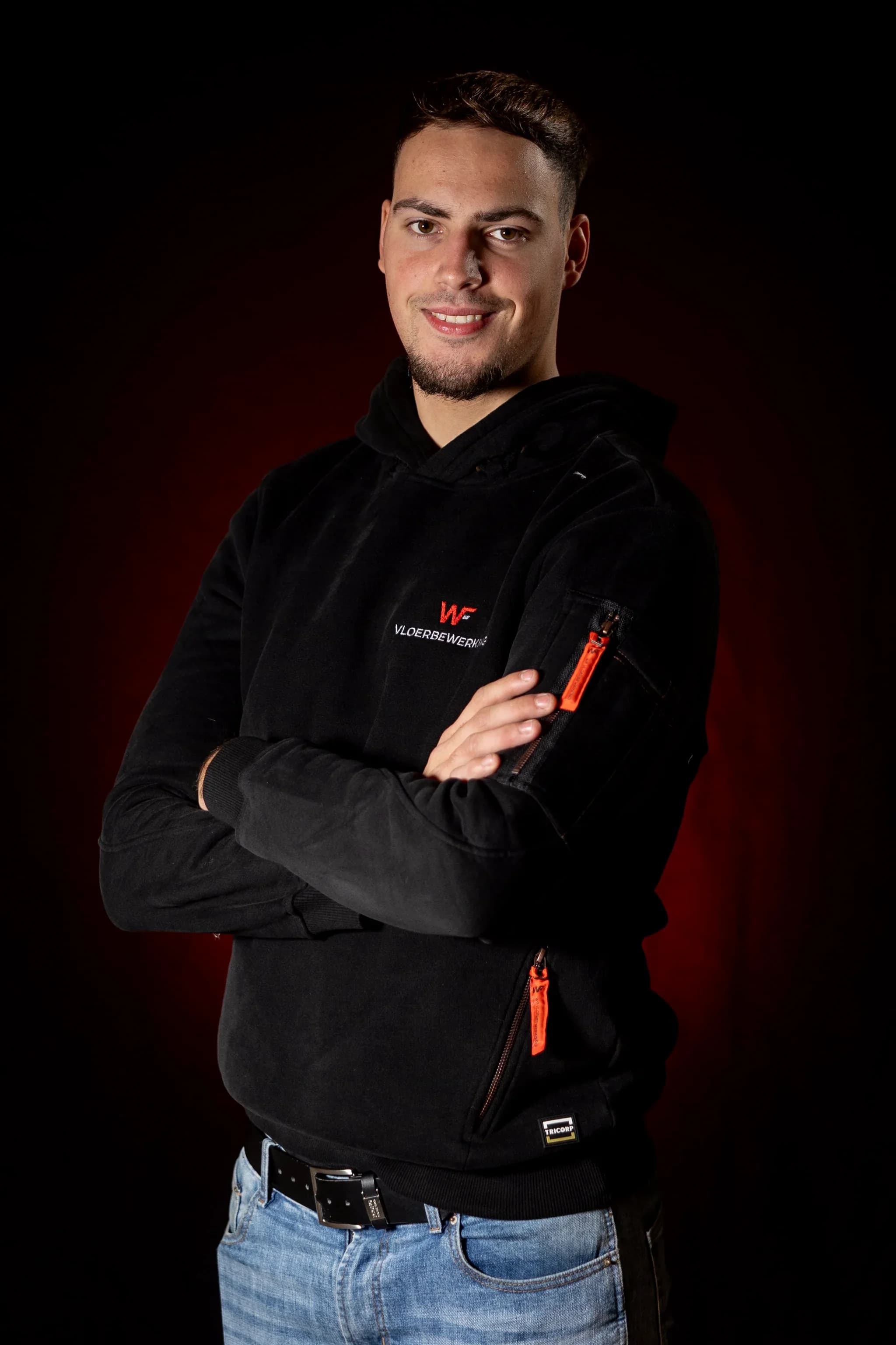 Jonge man poseert met gekruiste armen in zwarte hoodie met oranje ritsdetails en zichtbaar label 'TRICORP' tegen donkere achtergrond
