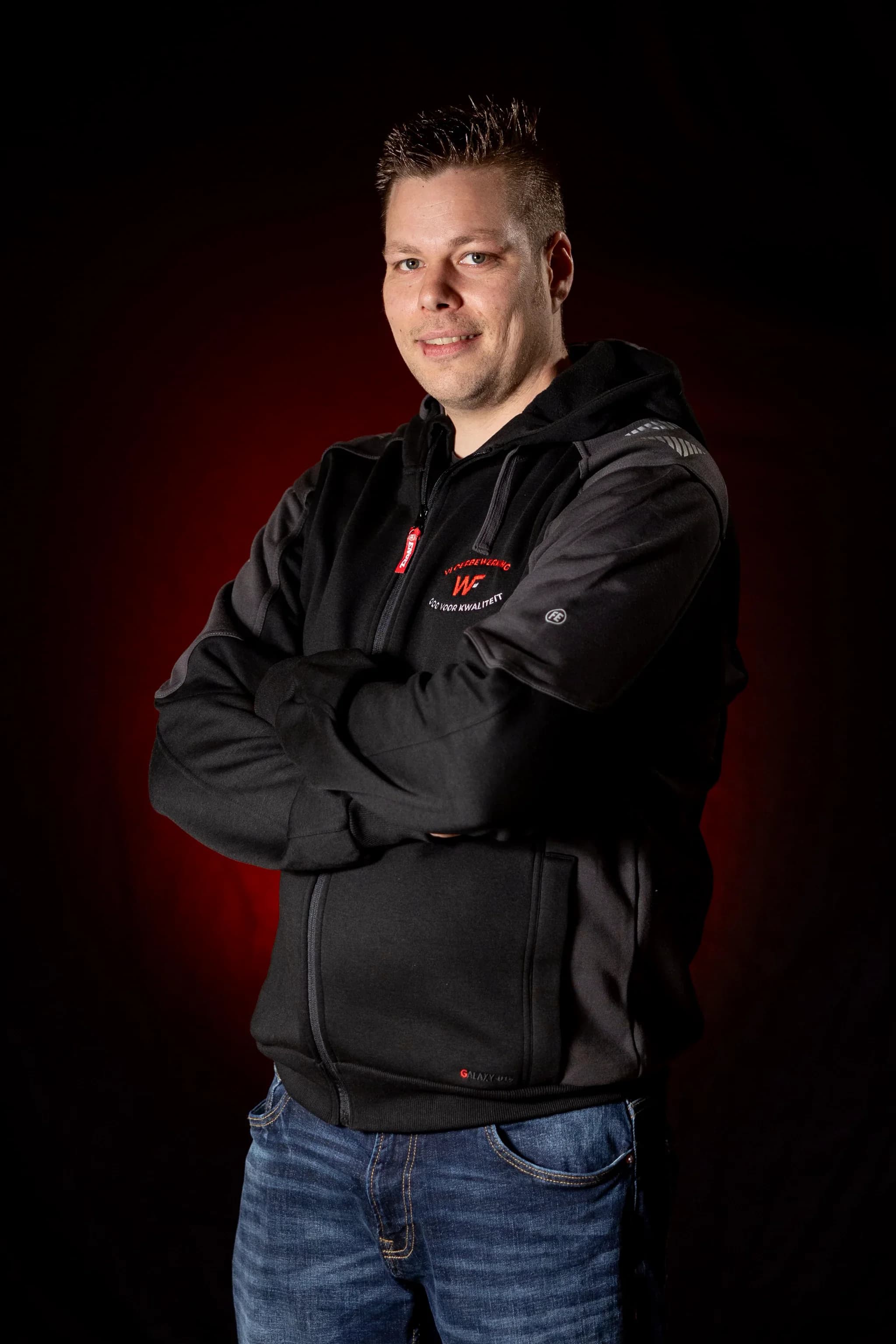 Portret van een man staand met gevouwen armen, hij draagt een zwart vest met capuchon en een blauwe spijkerbroek, op een donkere achtergrond met rode gloed.