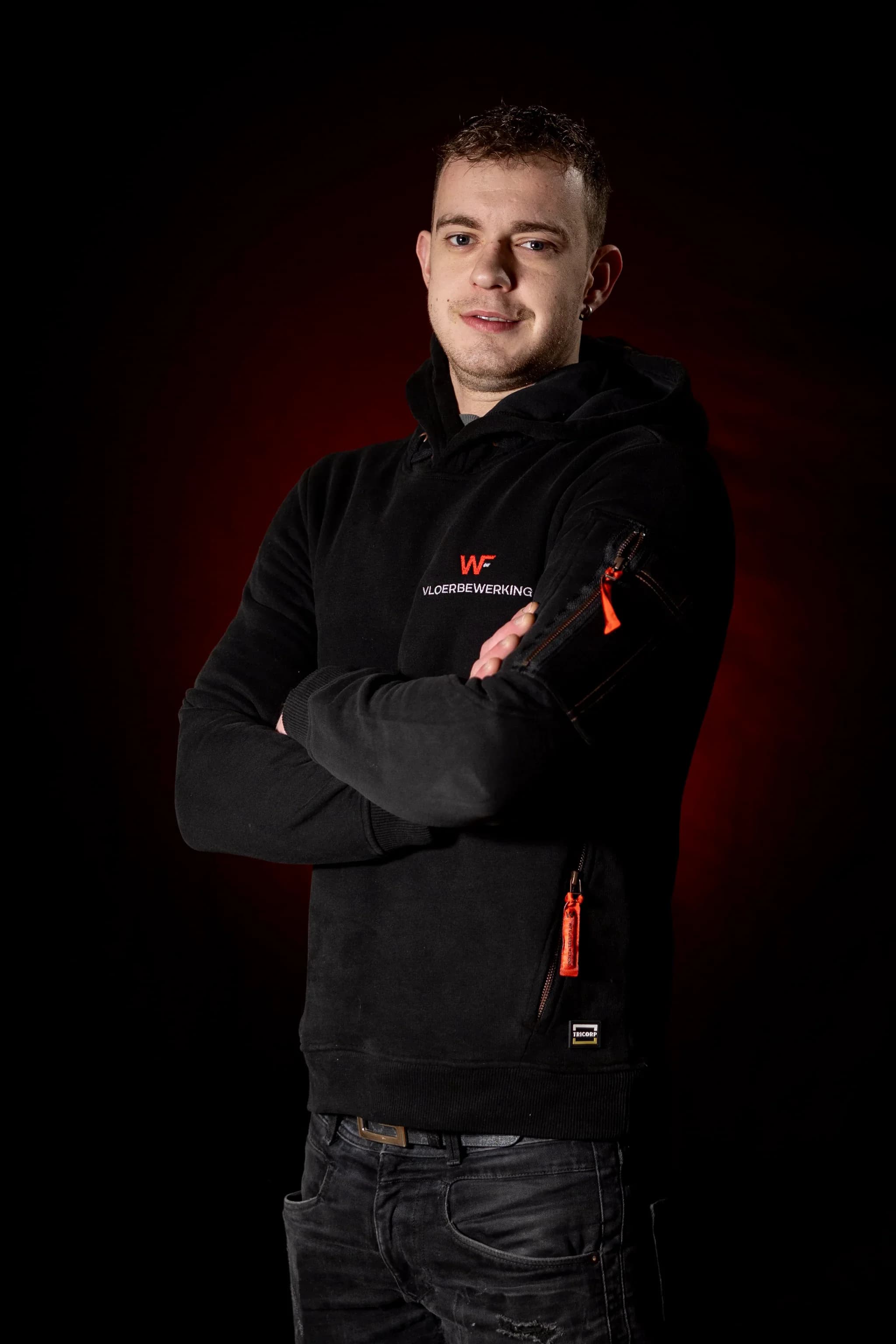 Portret van een man in zwarte hoodie met de tekst 'VLOERBEWERKING' en een rood WF-logo, armen gekruist en donkere achtergrond met rode gloed.