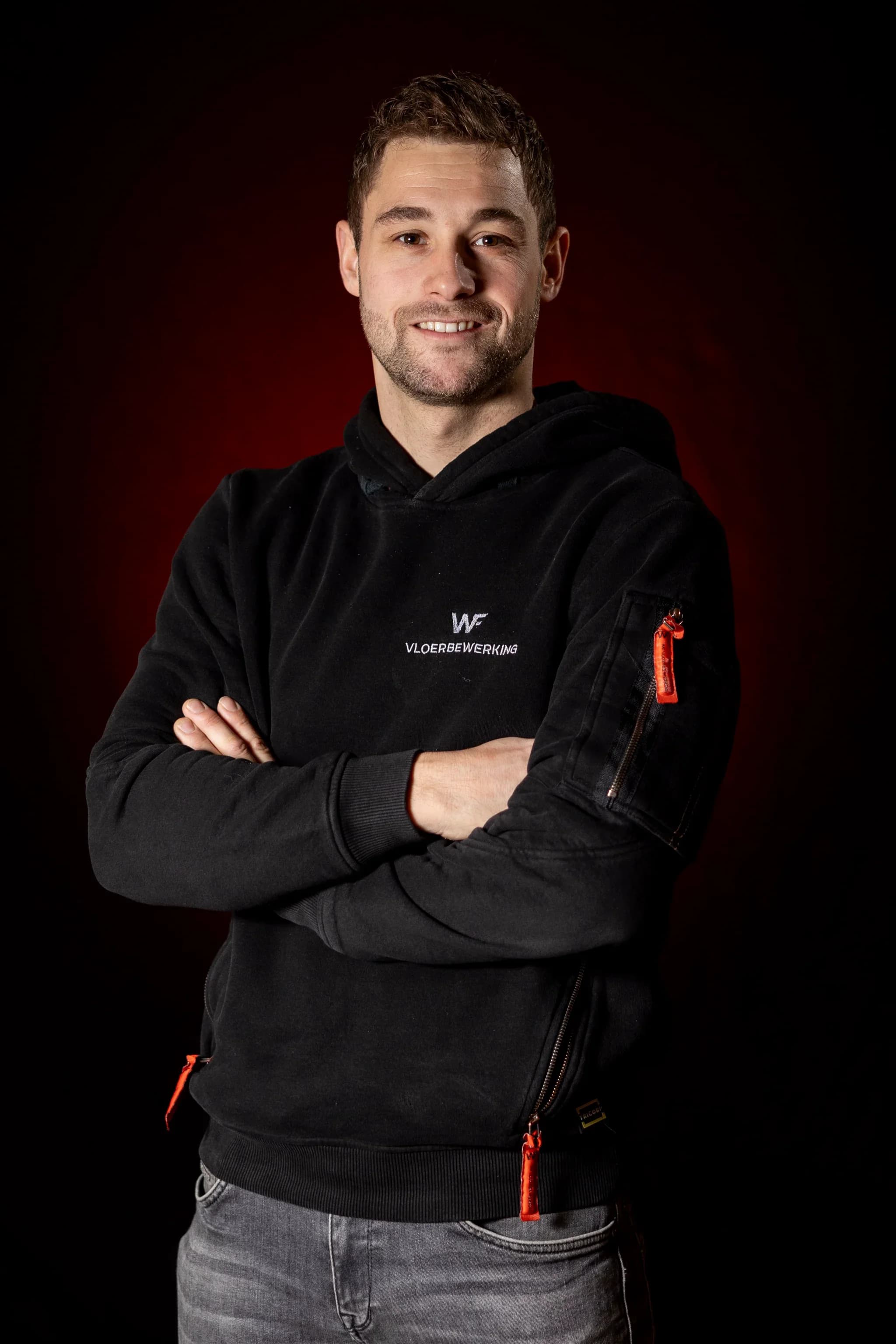 Halflang portret van een lachende man met gekruiste armen, zwarte hoodie met tekst 'VLOERBEWERKING', donkere roodachtige achtergrond.