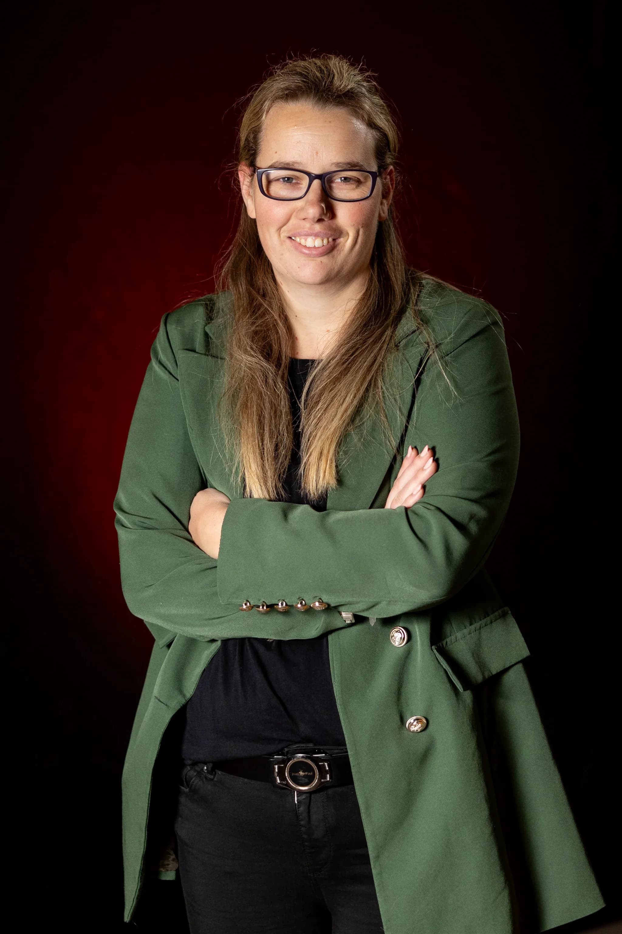 Portret van een vrouw met bril en lang haar, gekleed in een groen colbert en zwart shirt, met gekruiste armen voor een donkere achtergrond.