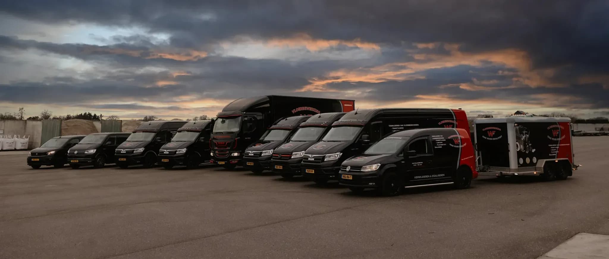 Zwarte bestelwagens en vrachtwagen naast elkaar op een parkeerterrein onder een bewolkte avondlucht met oranje en blauwe tinten.