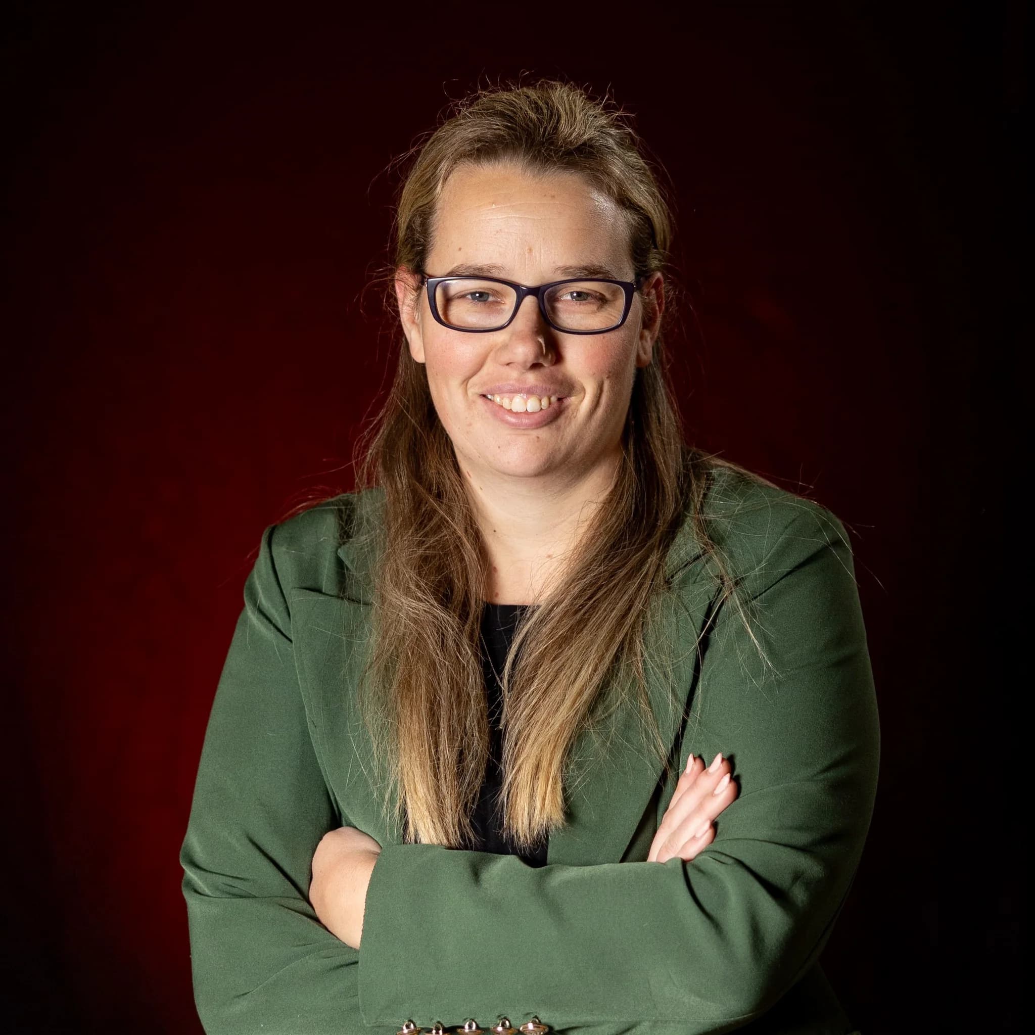 Lachend portret van een volwassen persoon met bril en groene blazer, armen gekruist, op donkere rode achtergrond