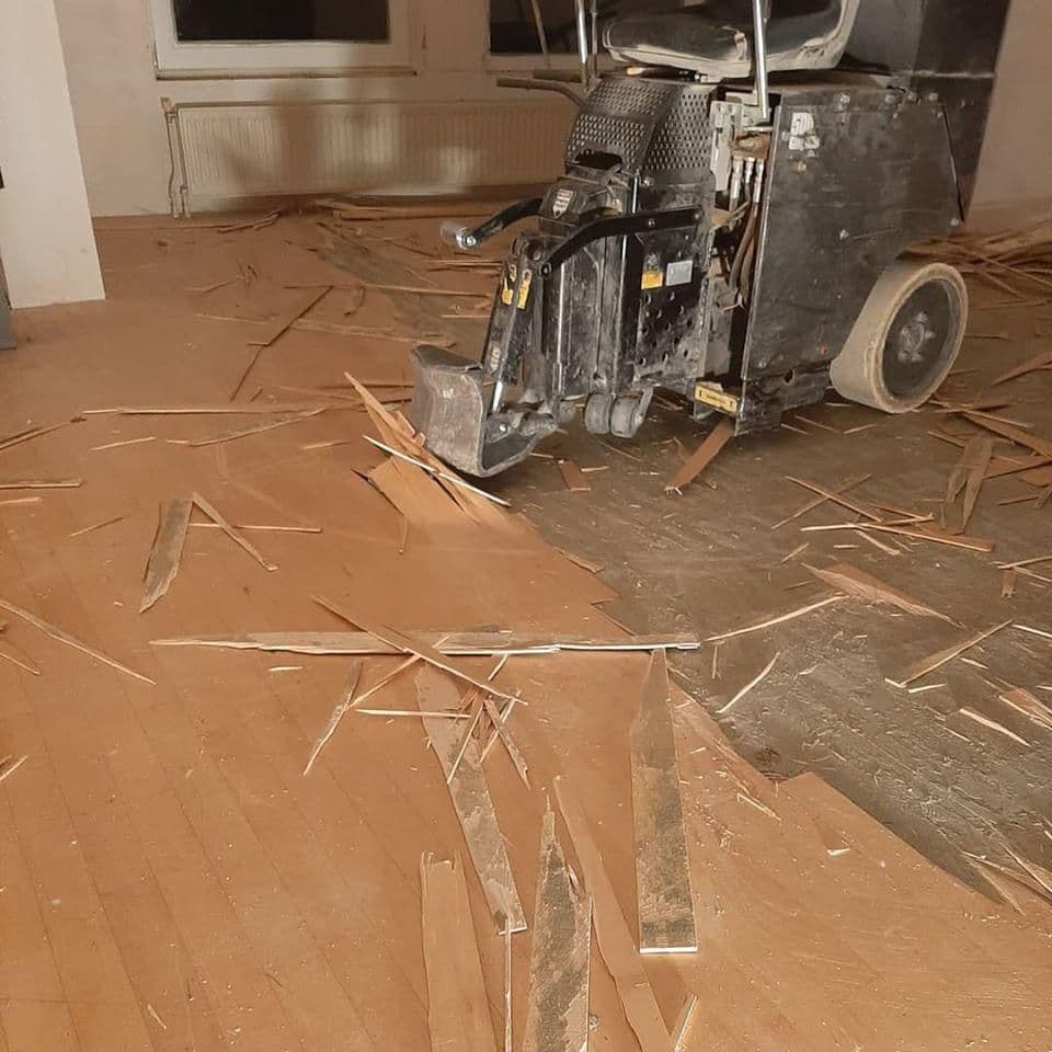 Zwarte vloerverwijderingsmachine in een kamer met deels verwijderde houten vloer en verspreide planken en splinters.