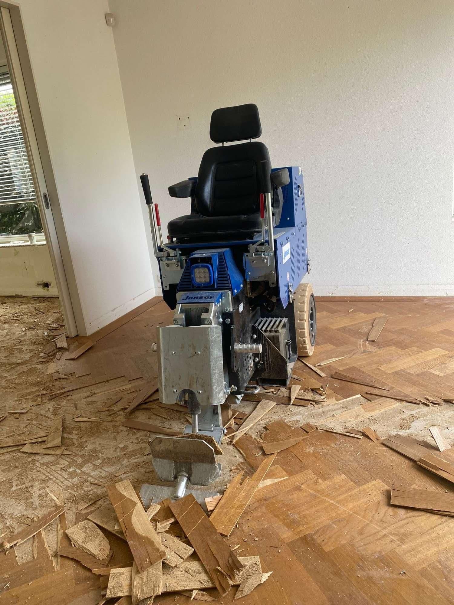 Blauwe machine met stoel die houten vloerpanelen verwijdert in een kamer met visgraatparket.