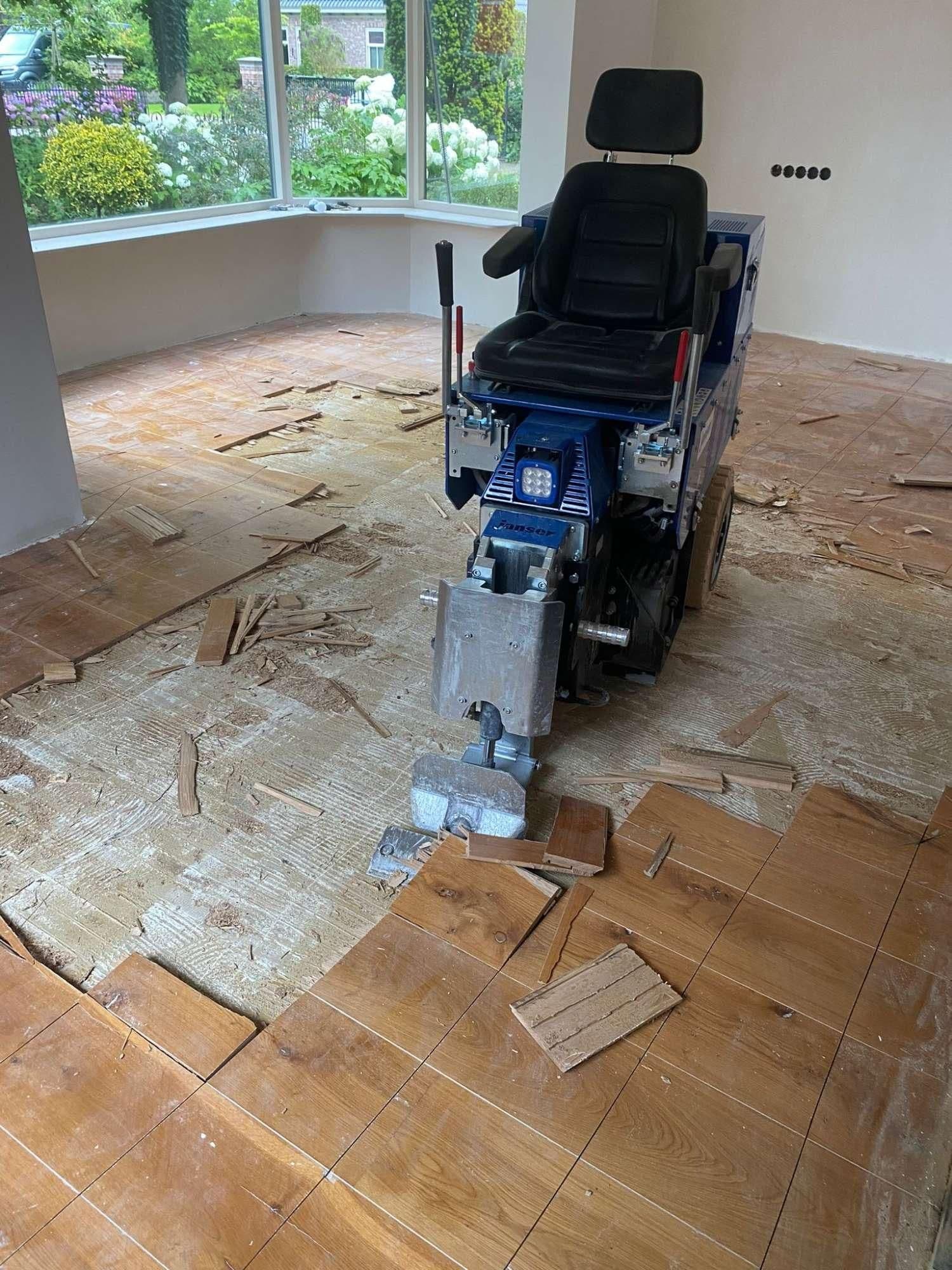 Machine voor het verwijderen van houten vloerdelen op een vloer in renovatie met grote ramen en buitenzicht op tuin.