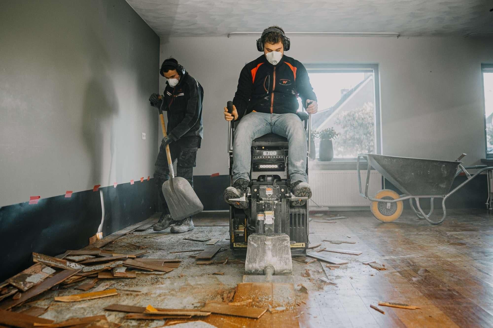 Twee mannen breken houten vloer af met machine en schop in een kamer