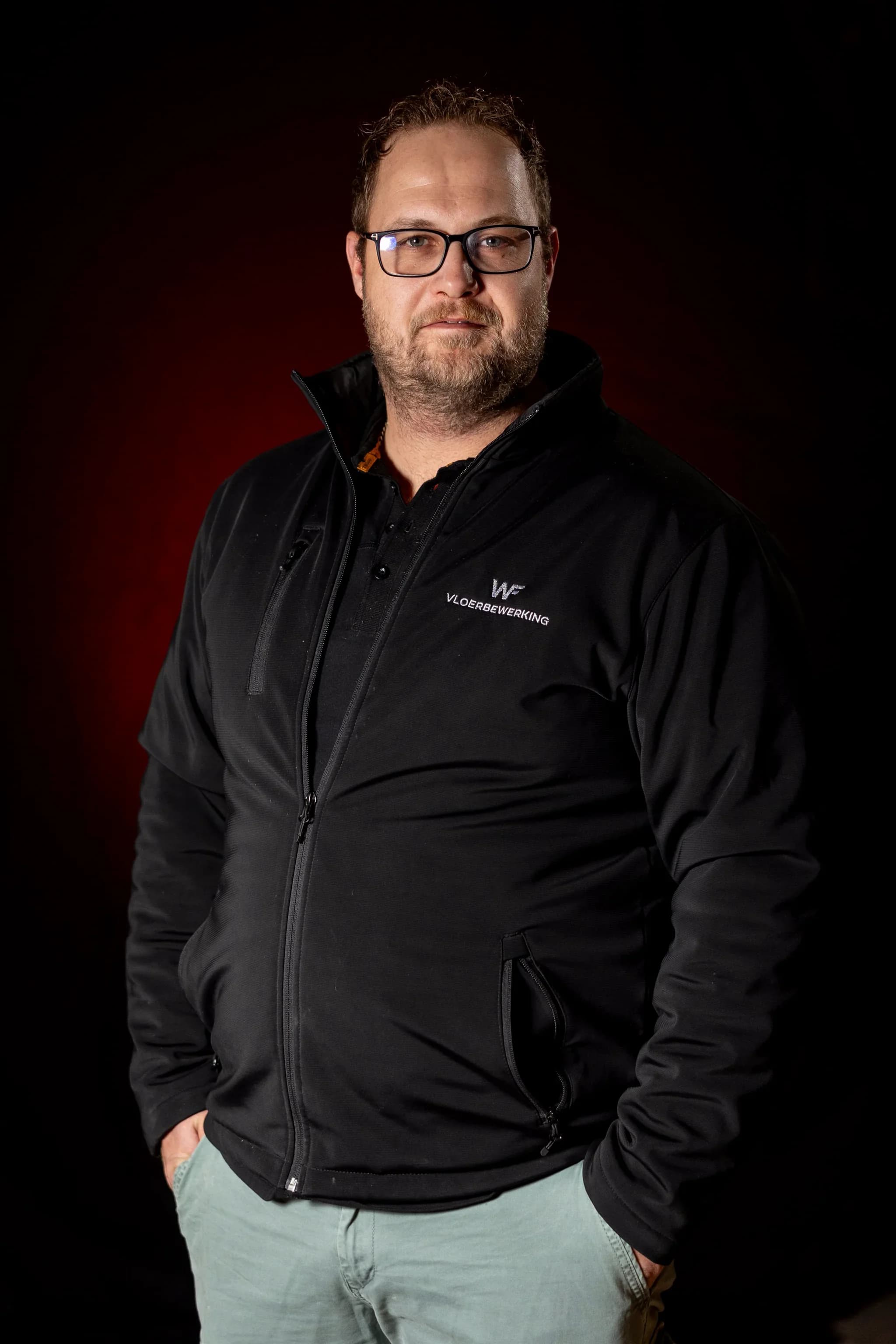 Portret van een man met bril en baard in een zwart jack met de tekst 'VLOERBEWERKING', handen in de zakken, tegen een donkere achtergrond.