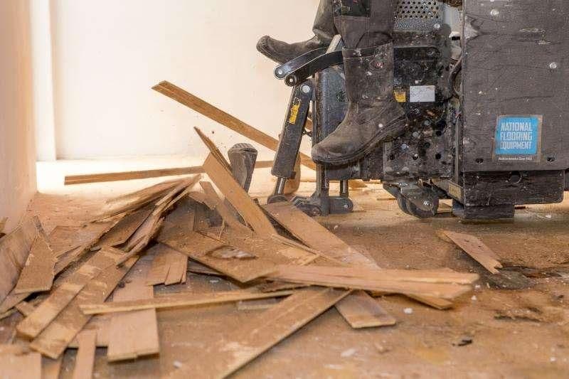 Persoon met laarzen gebruikt een vloerschuurmachine om houten vloer te schaven met afgepelde vloerdelen op de grond