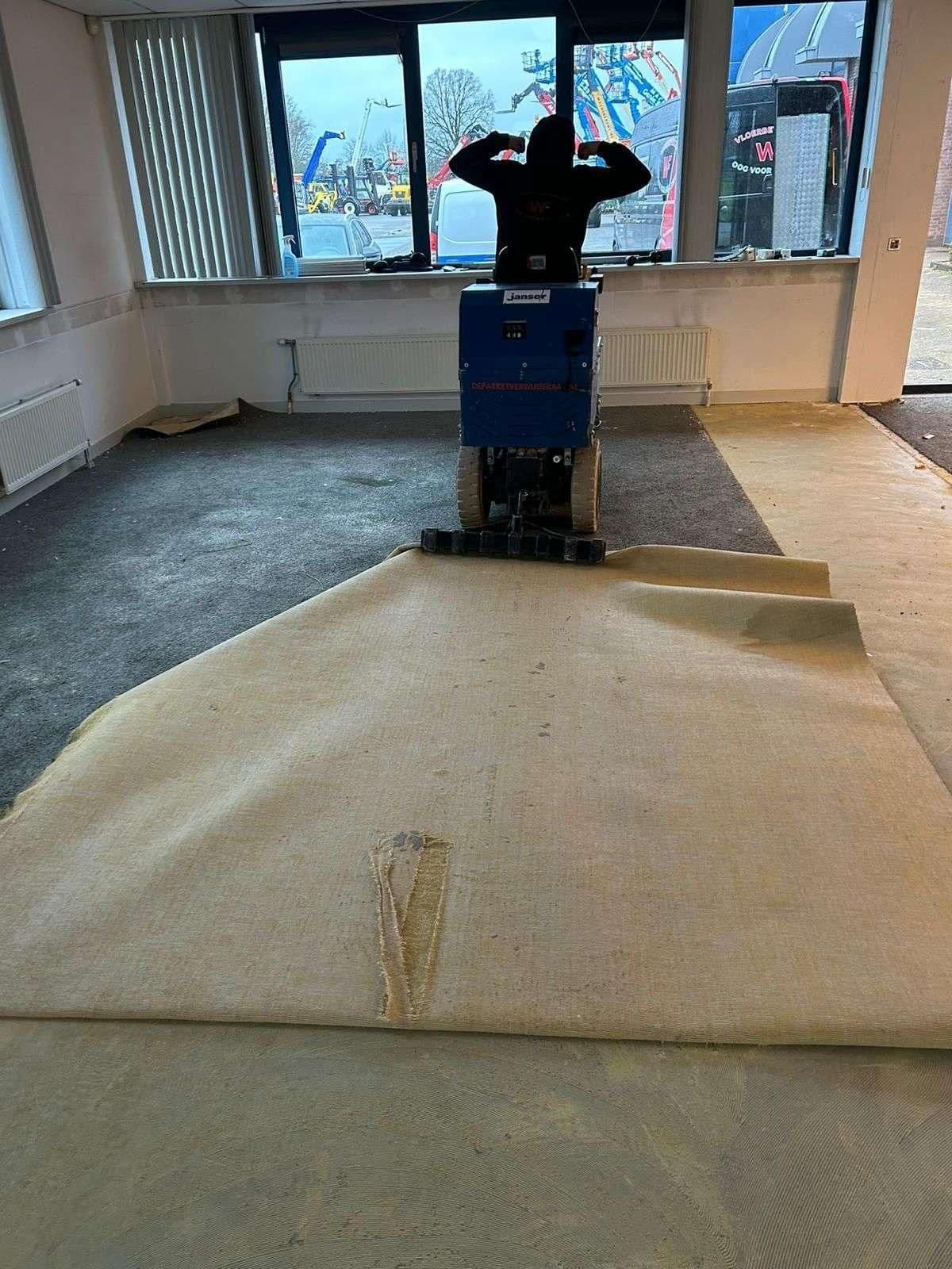 Persoon op een machine die vloerbedekking losmaakt in een binnenruimte met grote ramen