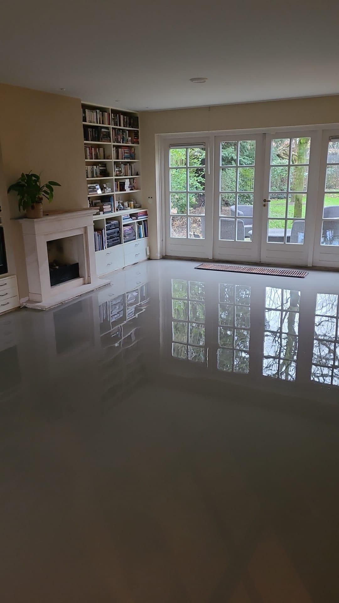 Woonkamer met glanzende reflecterende vloer, witte schouw, boekenkast en grote glazen deuren met zicht op de tuin.