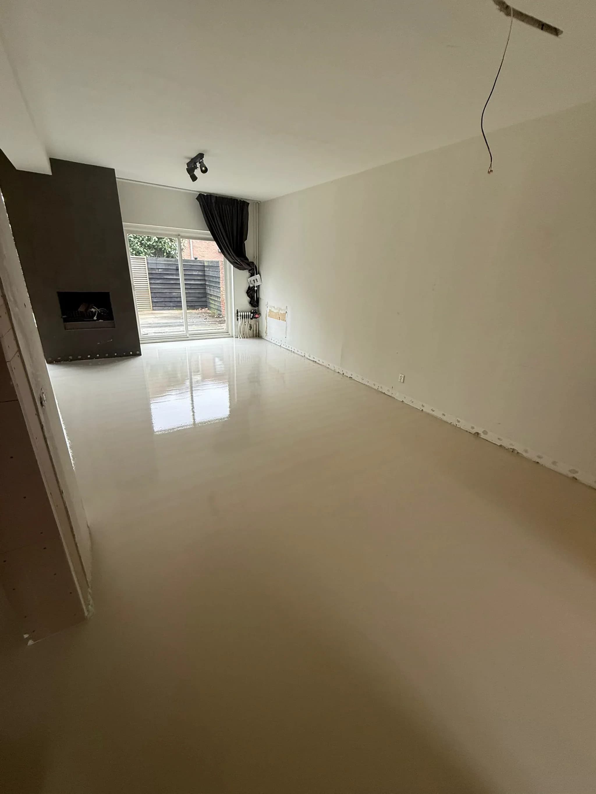 Lege woonkamer tijdens renovatie met glanzende lichte vloer, witte muren, haard links, schuifpui met donker gordijn en loshangende elektriciteitskabel
