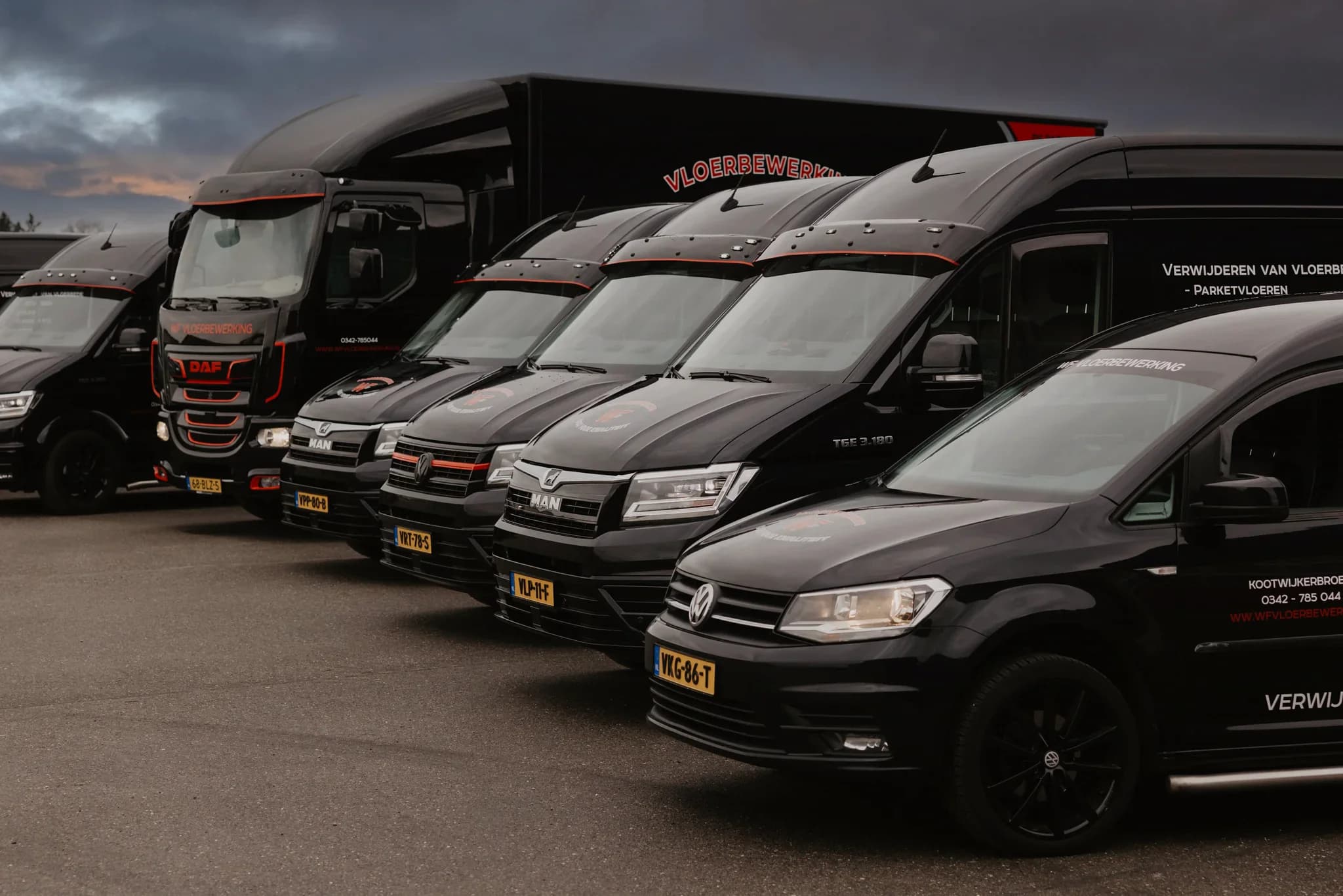 Rij zwarte bedrijfsbussen en een vrachtwagen met bedrijfstekst 'VLOERBEWERKING' geparkeerd op een parkeerterrein