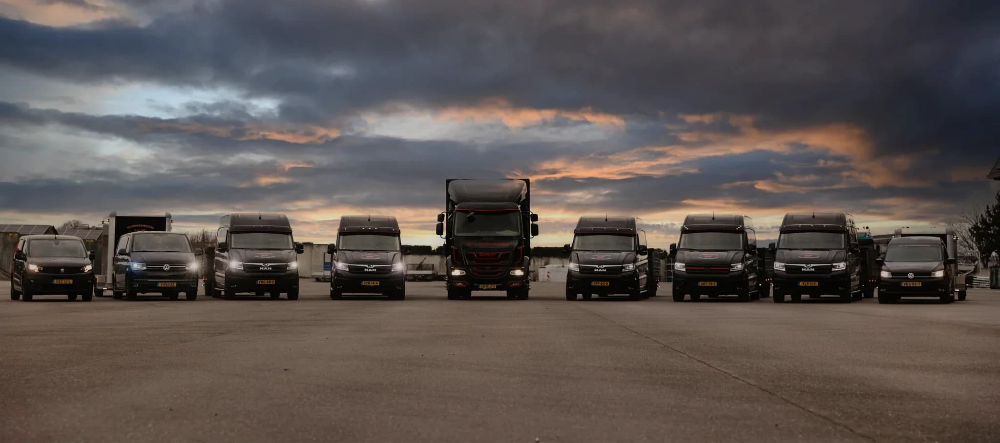 Zwarte bestelwagens en een vrachtwagen frontaal geparkeerd op een open terrein onder een bewolkte avondlucht met zachte kleuren.